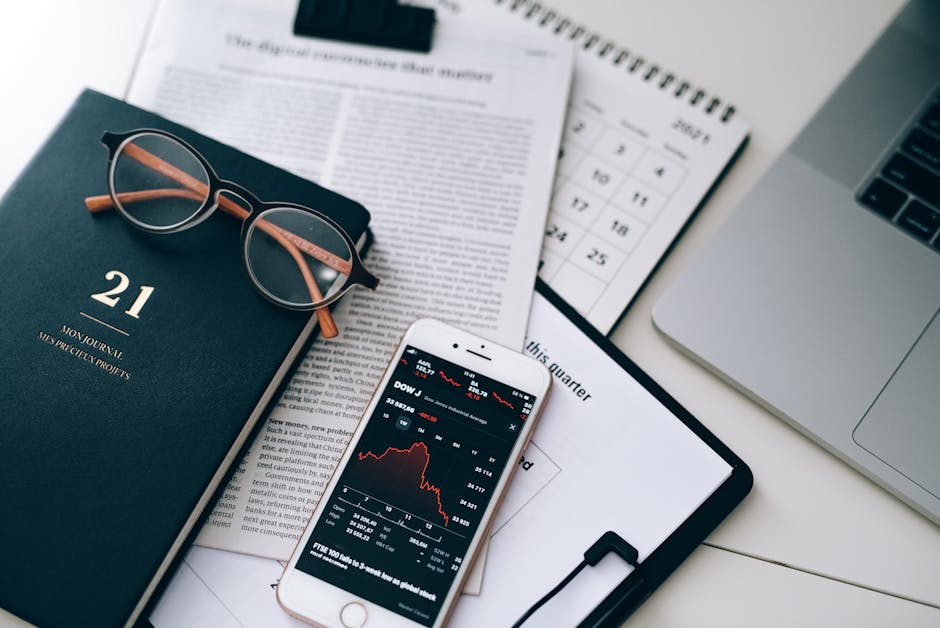 A stylish workspace featuring financial documents, eyeglasses, an iPhone displaying stock data, and a laptop.